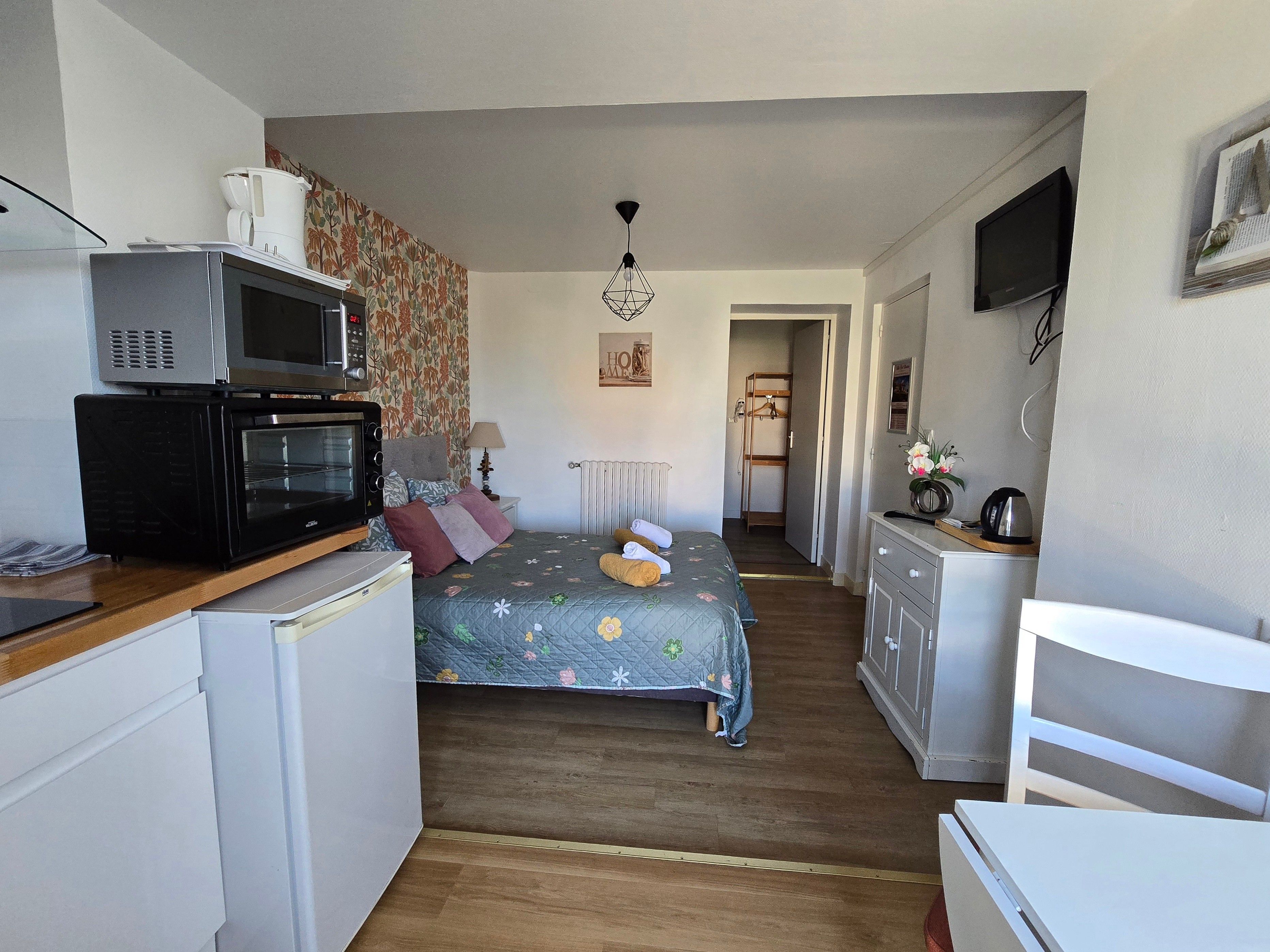 A compact hotel room with a kitchenette, a bed with floral bedding, a small desk, and a television.