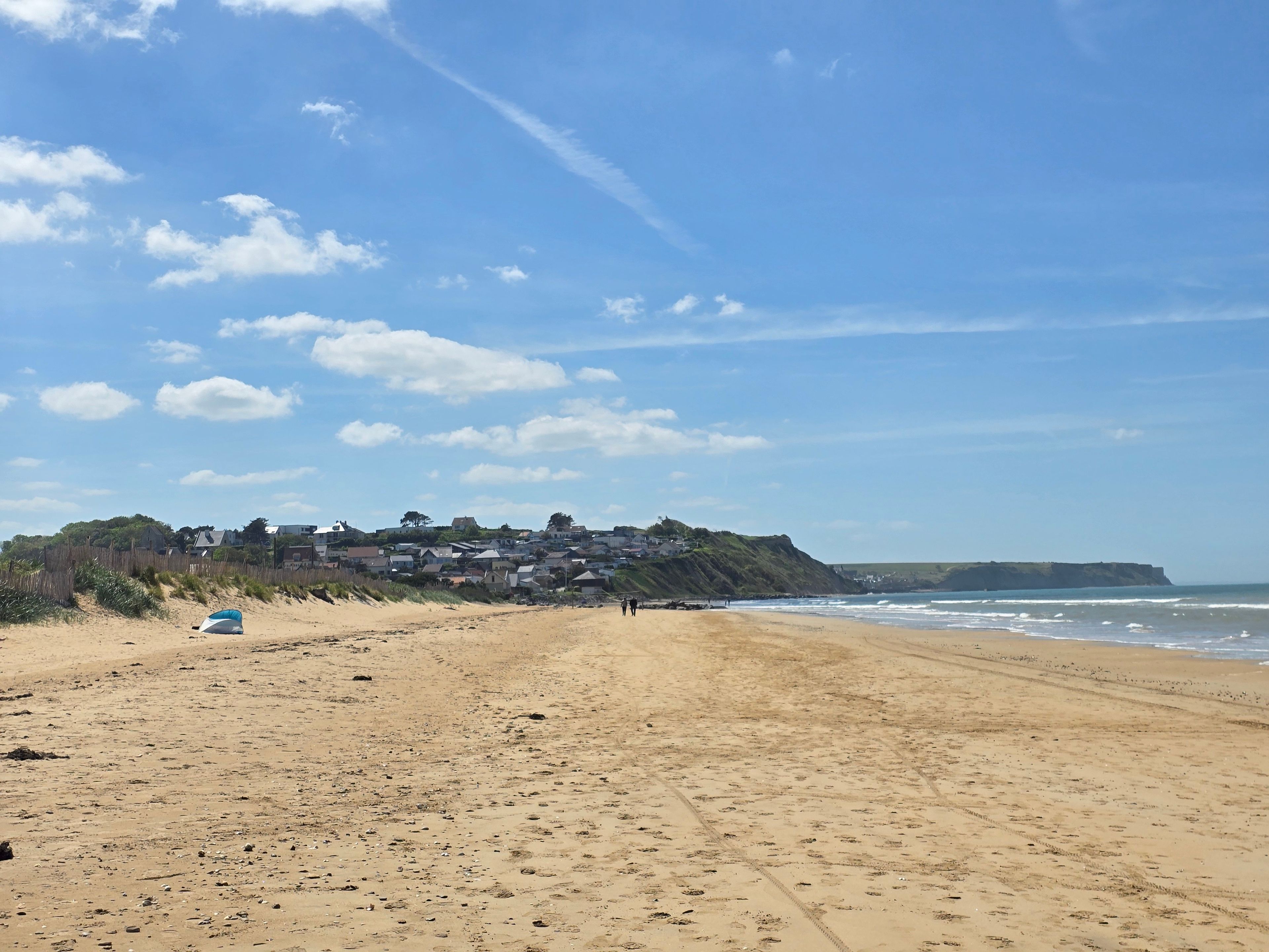 A sandy beach with a clear sky, a few scattered clouds, and a distant view of houses on a hill.