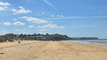 A sandy beach with a clear sky, a few scattered clouds, and a distant view of houses on a hill.