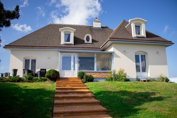 A two-story house with a front porch, a chimney, and a large window. - Studio 'Charmant Bord De Mer' with Private Terrace, Shared Garden and Wi-Fi (Saint-Côme-de-Fresné)