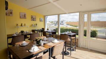 A dining area with tables set for four, a yellow wall with artwork, and a view of a field through large windows.