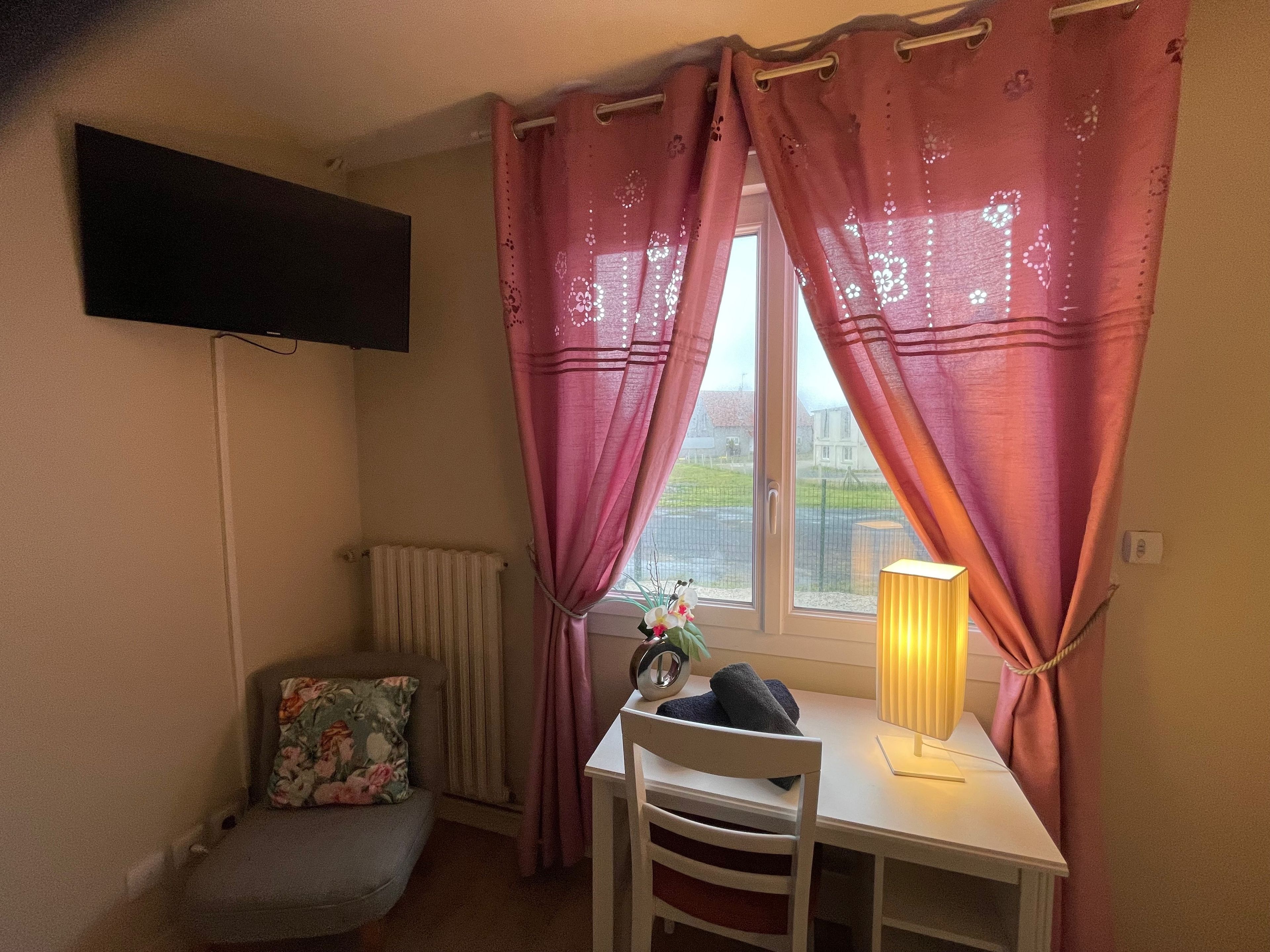 A small room with a single table, a chair, a radiator, a TV mounted on the wall, and a window with pink curtains.