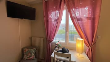 A small room with a single table, a chair, a radiator, a TV mounted on the wall, and a window with pink curtains.