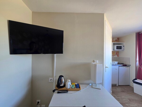 A room with a flat-screen TV mounted on the wall, a small kitchenette with a microwave, and a table with a kettle and a tray of items. - Studio 'La Parenthèse Normande - Terrace' for 2 People with Private Terrace and Wi-Fi (Saint-Côme-de-Fresné)