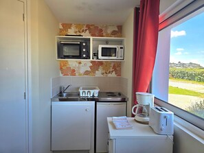A compact kitchenette with a microwave, toaster, and coffee maker on a small table, and a window with a view of greenery. - Studio 'La Parenthèse Normande - Terrace' for 2 People with Private Terrace and Wi-Fi (Saint-Côme-de-Fresné)