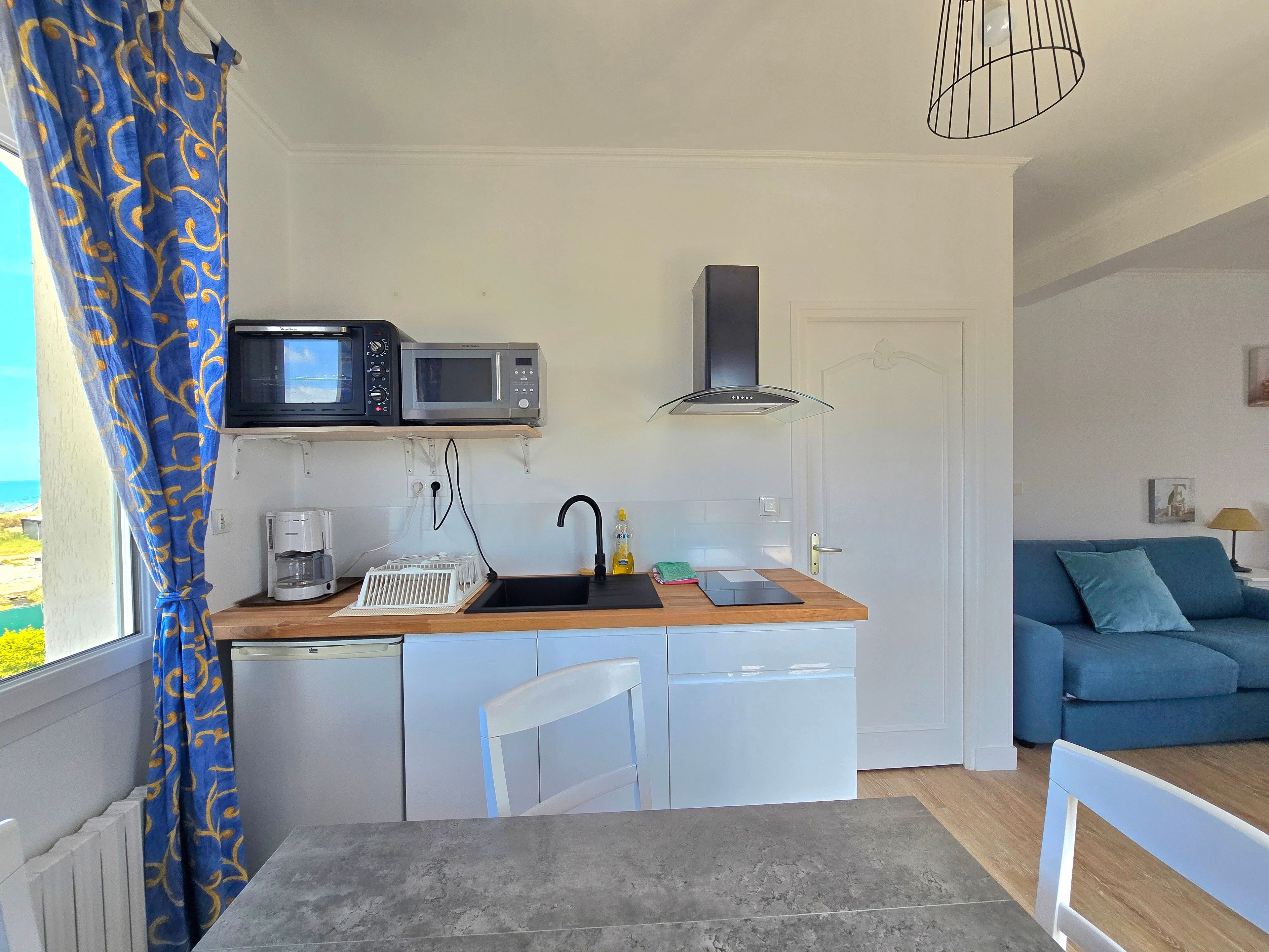 Cocina compacta con microondas, pileta y cafetera. Hay un comedor con mesa y sillas, y un sofá azul en la sala.