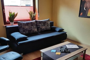 Ein modernes Wohnzimmer mit einer schwarzen Couch, zwei gemusterten Kissen, einem Couchtisch mit Gegenständen und zwei Topfpflanzen.