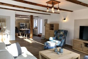 A living room with a sofa, ottoman, and a coffee table. There is a television on a wooden cabinet, and a patterned armchair. A dining area with a table and chairs is visible in the background. - Mountain apart 5||Wernigerode (Wernigerode)