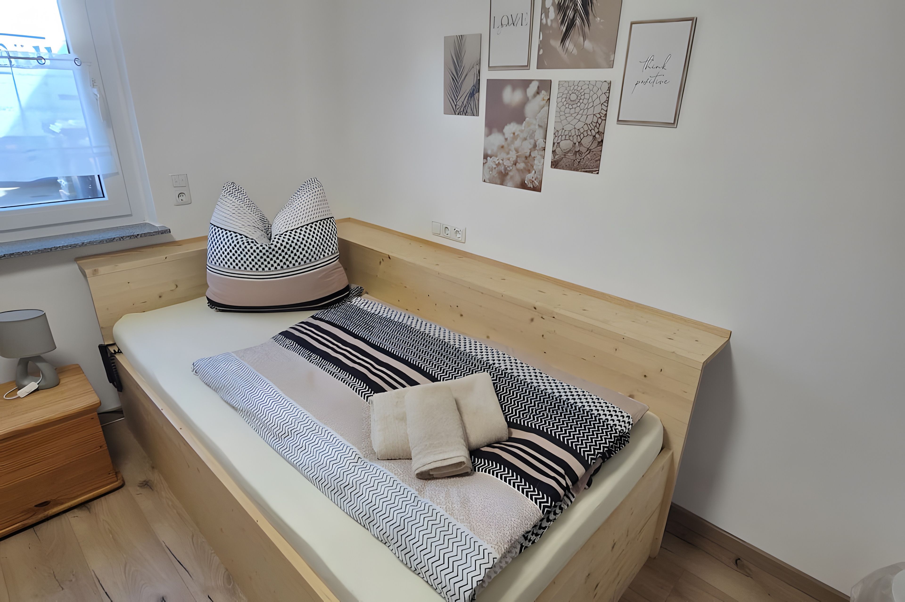 Ein kleines, ordentlich bezogenes Bett mit einem Holzkopfteil, ein Nachttisch mit Lampe und eine Wand mit gerahmten Bildern.