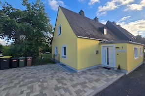 Ein gelbes Gebäude mit einem geneigten Dach, einer Tür mit der Aufschrift 'Drosselweg' und einem gepflasterten Eingangsbereich.