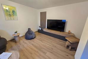 Una sala moderna con un televisor de pantalla plana, un sillón de peluche, una mesita auxiliar con una planta y un cuadro en la pared.