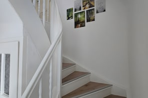 Un escalier avec des rampes blanches et des marches en bois mène à un palier où sont accrochées des images aux murs.