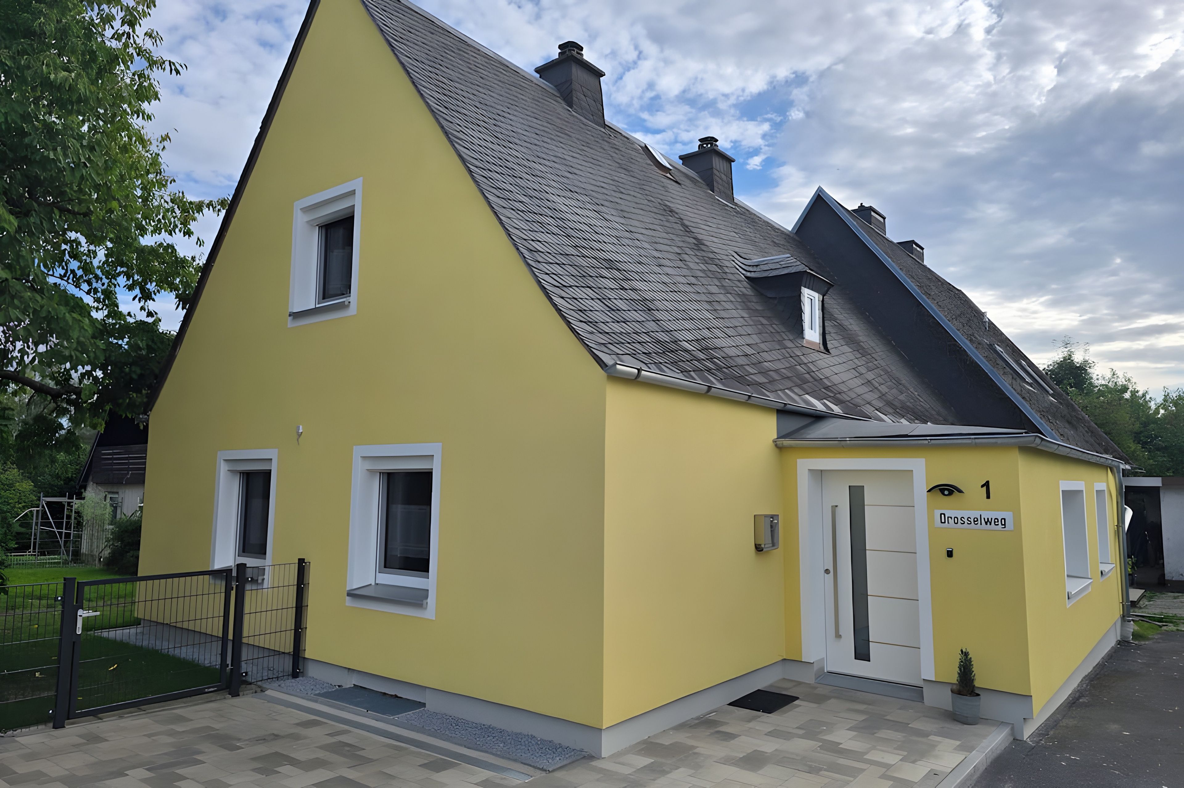 Ein gelbes Gebäude mit einem steilen Dach, einer Tür mit der Aufschrift 'Drosselweg' und einem kleinen Fenster.