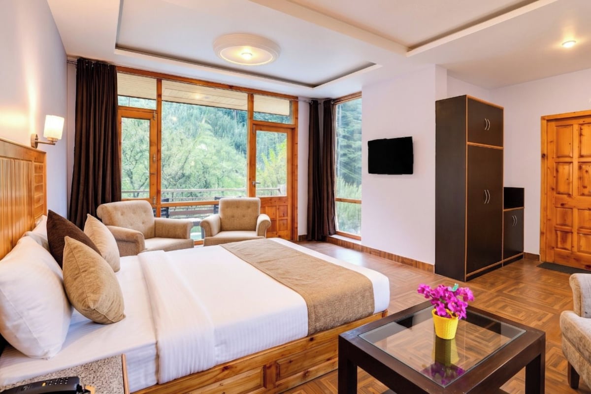 Una habitación de hotel con una cama grande, dos sillones, una mesa de centro con flores, un televisor y un armario.