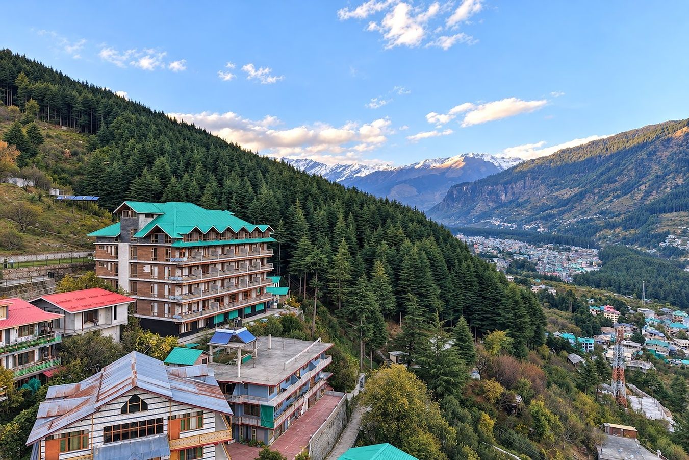 A multi-story hotel with a green roof, surrounded by dense forest and mountains in the background.