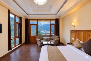 Habitación de hotel con una cama grande, un sofá, una mesita y vistas a las montañas a través de amplios ventanales.