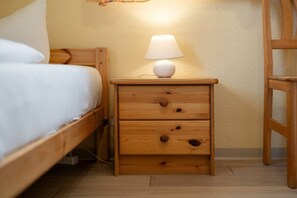 Uma mesa de cabeceira de madeira com uma lâmpada, uma cama com roupa de cama branca e uma cadeira de madeira.