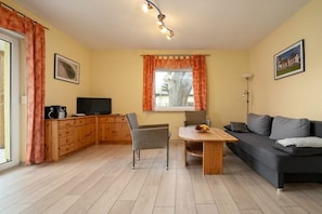 Een woonkamer met een bank, een eettafel, een televisie en een keuken met kasten en een raam.