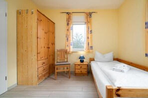 A bedroom with a bed, a wooden wardrobe, a chair, a nightstand with a lamp, and a window with curtains. - Holiday apartment Haus Vietzen 2 (Rechlin)