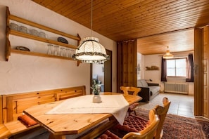 A wooden dining table with benches, a chandelier, and a view into a living area with a sofa and a window. - Holiday apartment with 1 bedroom (Füssen-Hopfen am See)