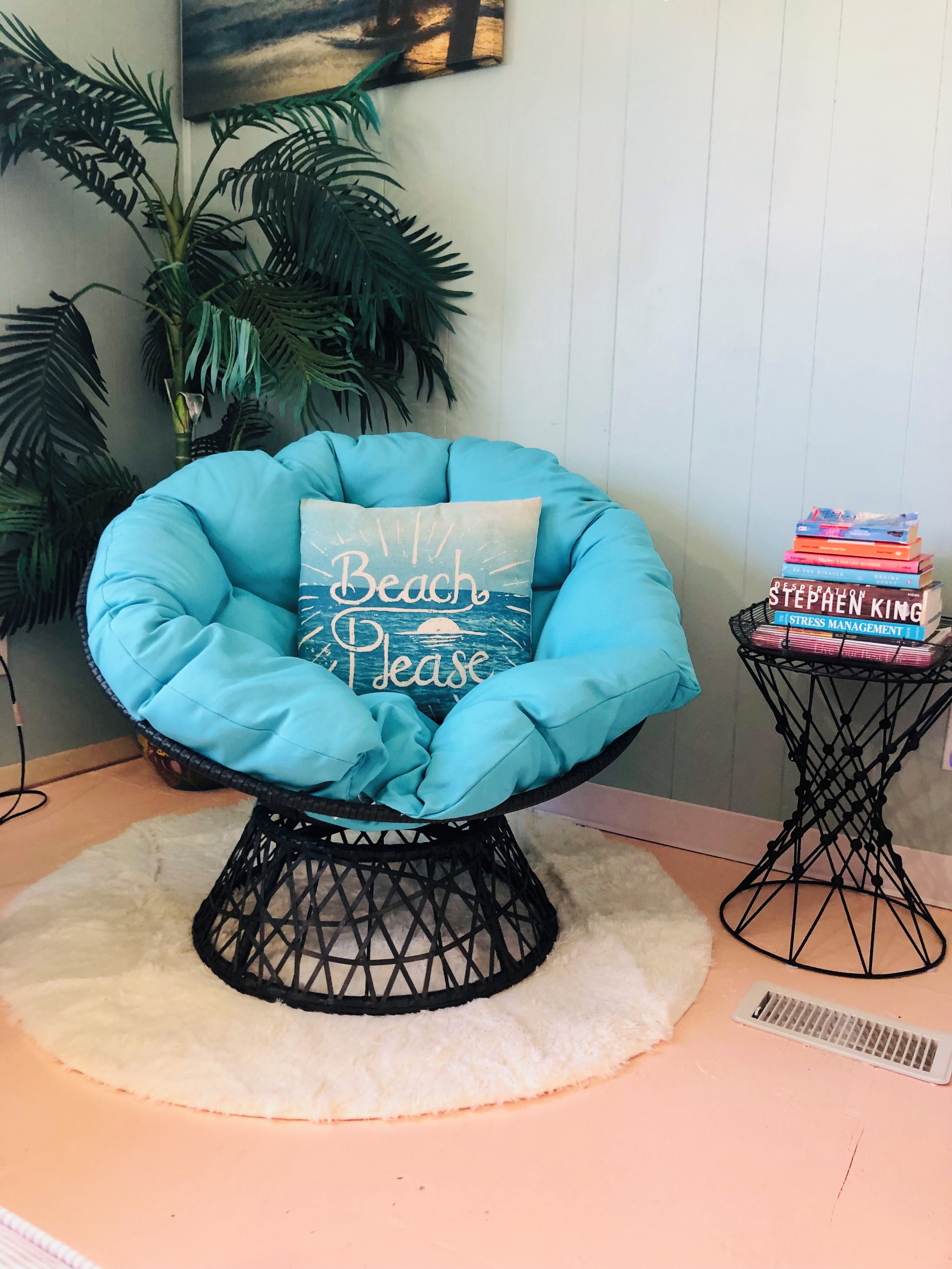 A blue chair with a cushion that reads 'Beach Please' and a stack of books on a side table.