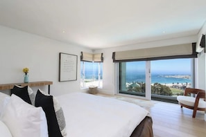 A bedroom with a large bed, a view of the sea, and a chair by the window. - 26 Geneva Drive - Sandpiper House (Cape Town)