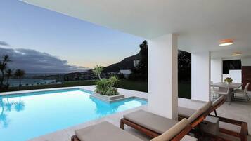 Un espace piscine extérieur moderne, doté de chaises longues, d’une table à manger et offrant une vue sur l’océan et les collines.