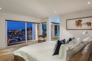 Een slaapkamer met een groot bed, uitzicht op de stad doorgaande ramen van vloer tot plafond, een lijst met olifanten en een nachtkastje met een lamp.