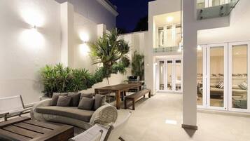 Un espace extérieur aménagé en terrasse, avec un canapé, une table à manger et des chaises, entouré de plantes et agrémenté d’un escalier menant à un niveau supérieur.