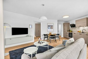 A modern living room with a sofa, a dining table, and a kitchen area with cabinets and a TV. - Holiday apartment with 1 bedroom (Dagebüll)