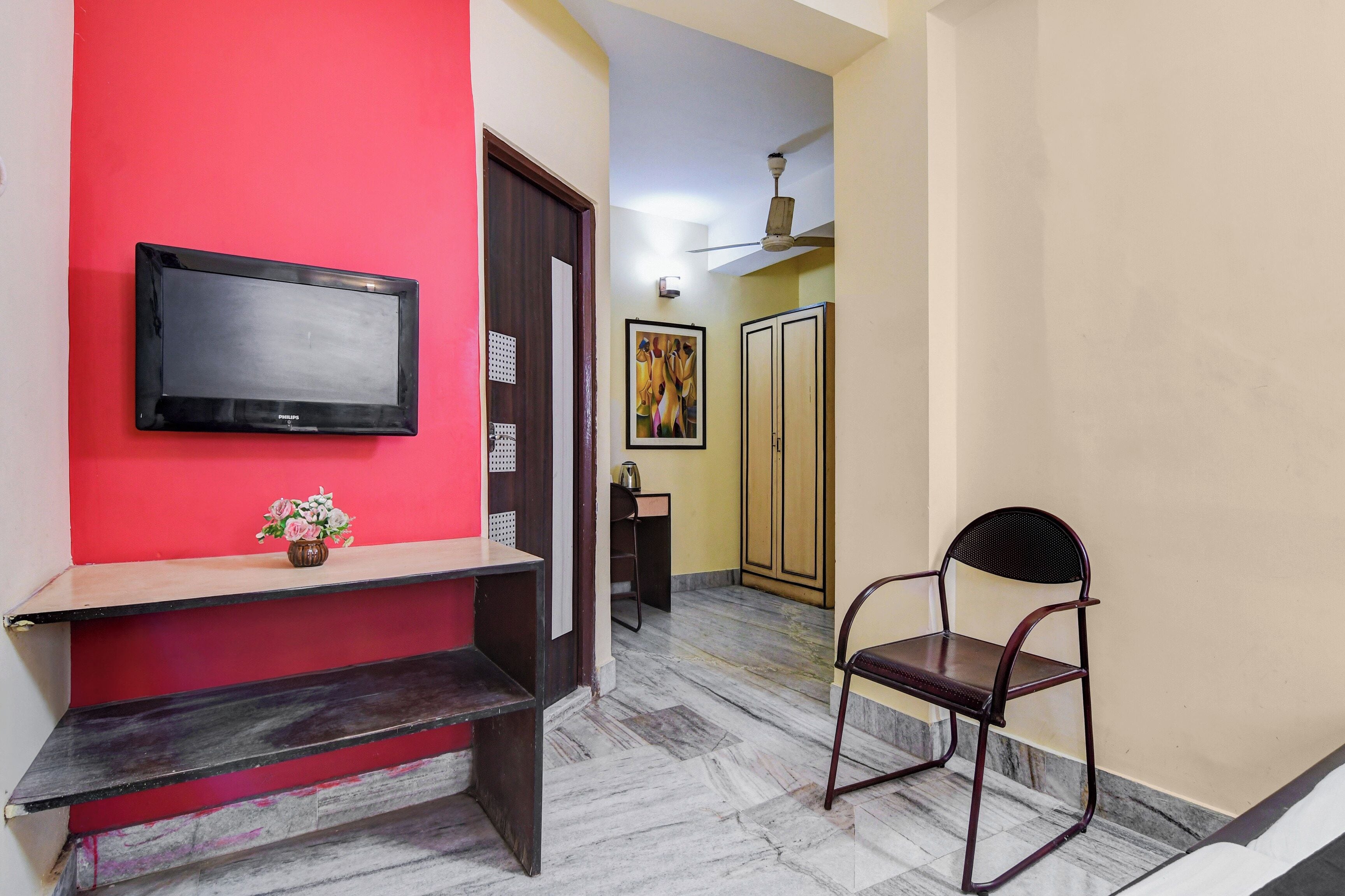 Un pasillo con un televisor de pantalla plana montado en una pared roja, un armario de madera, una silla y una puerta que da a otra habitación.