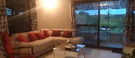 A living room with a sofa, chairs, a coffee table, and a view through a glass door to the outdoors.