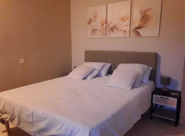 Ein ordentlich bezogenes Bett mit weißen Leinentüchern, ein Nachttisch mit Lampe und ein an der Wand hängendes Kunstwerk.