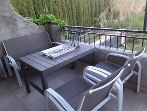 An outdoor patio with a table and chairs, a magazine on the table, and a view of greenery and a fence in the background.