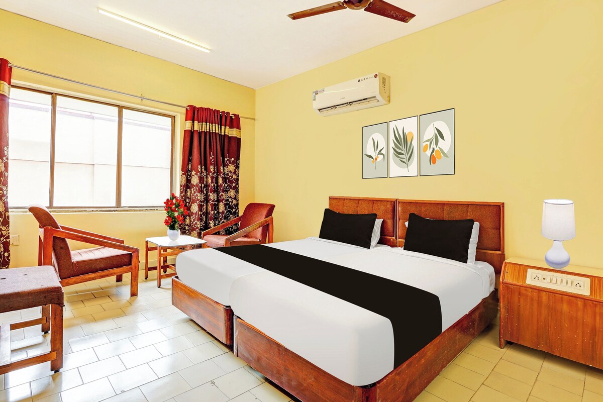 A hotel room with a bed, a desk, a chair, an air conditioning unit, and a wall-mounted lamp.