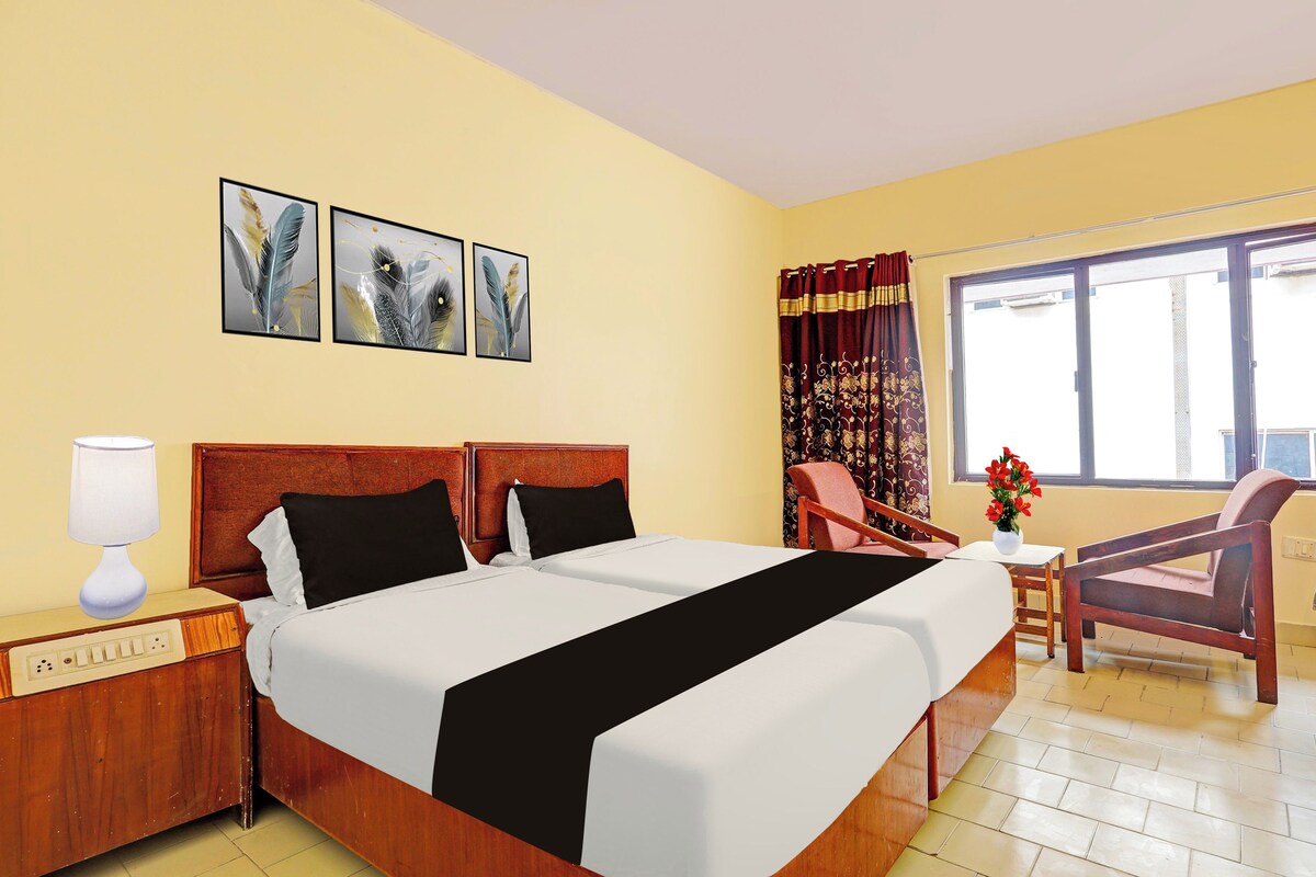 A hotel room with a double bed, a bedside table with a lamp, a chair, a small table with a vase, and a window with curtains.