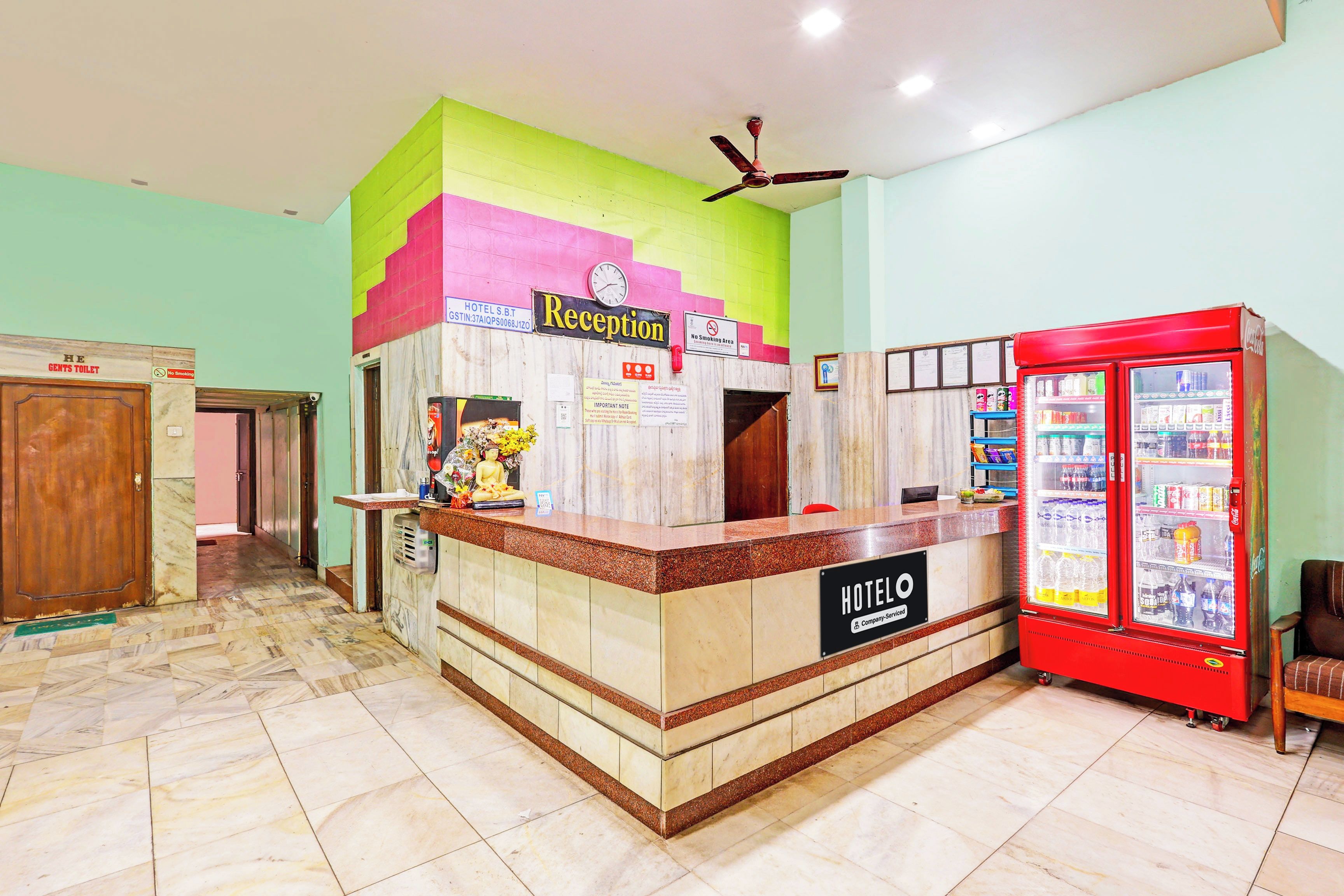 Un espace d’accueil d’hôtel comprenant un comptoir de réception, une horloge, un panneau « interdiction de fumer » et un réfrigérateur contenant des boissons Coca-Cola.