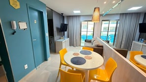 A modern hotel room with a dining area featuring a round table and yellow chairs, a TV mounted on the wall, and a view of the outside through large windows. - New Listing (Natal)