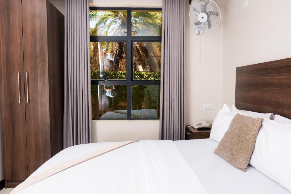 Habitación de hotel con cama, mesita de noche, ventilador, ventana con vista a follaje y armario de madera.