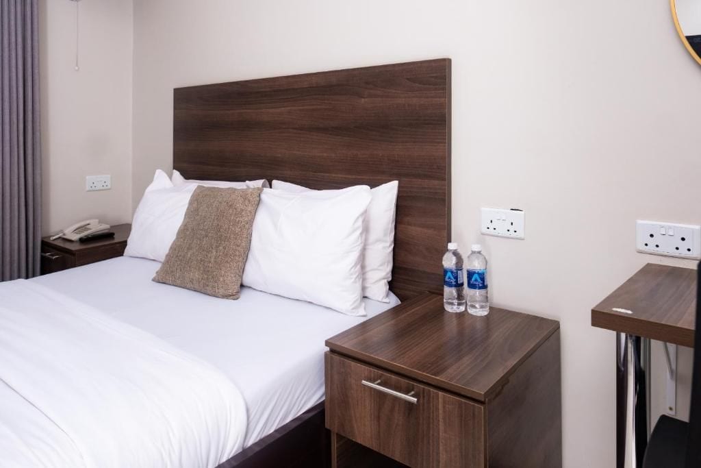 Habitación de hotel con cabecera de madera, cama con ropa de cama blanca, mesita de noche con un teléfono y una botella de agua, y una pequeña mesa de madera.
