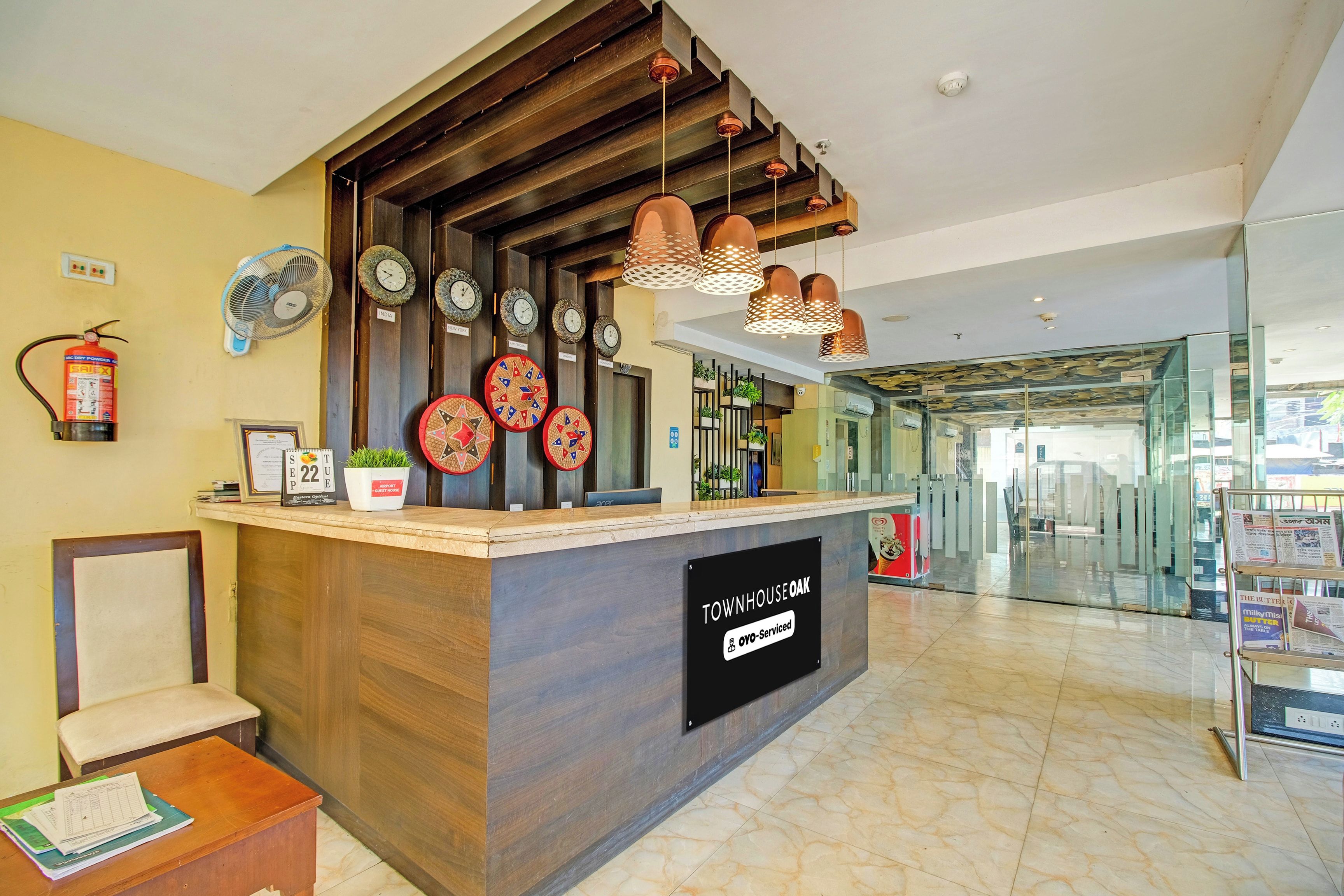 A reception desk with a sign indicating it is serviced by OYO. There are clocks on the wall, a fire extinguisher, and a fan.