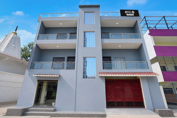 Un hotel de varios pisos con una puerta de entrada roja destacada, balcones en cada planta y un cielo despejado al fondo.