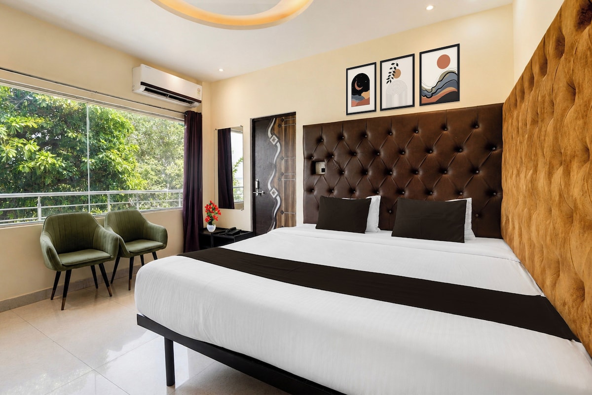 Ein modernes Hotelzimmer mit einem großen Bett, zwei grünen Sesseln, Blick ins Grüne und Kunstwerken an der Wand.