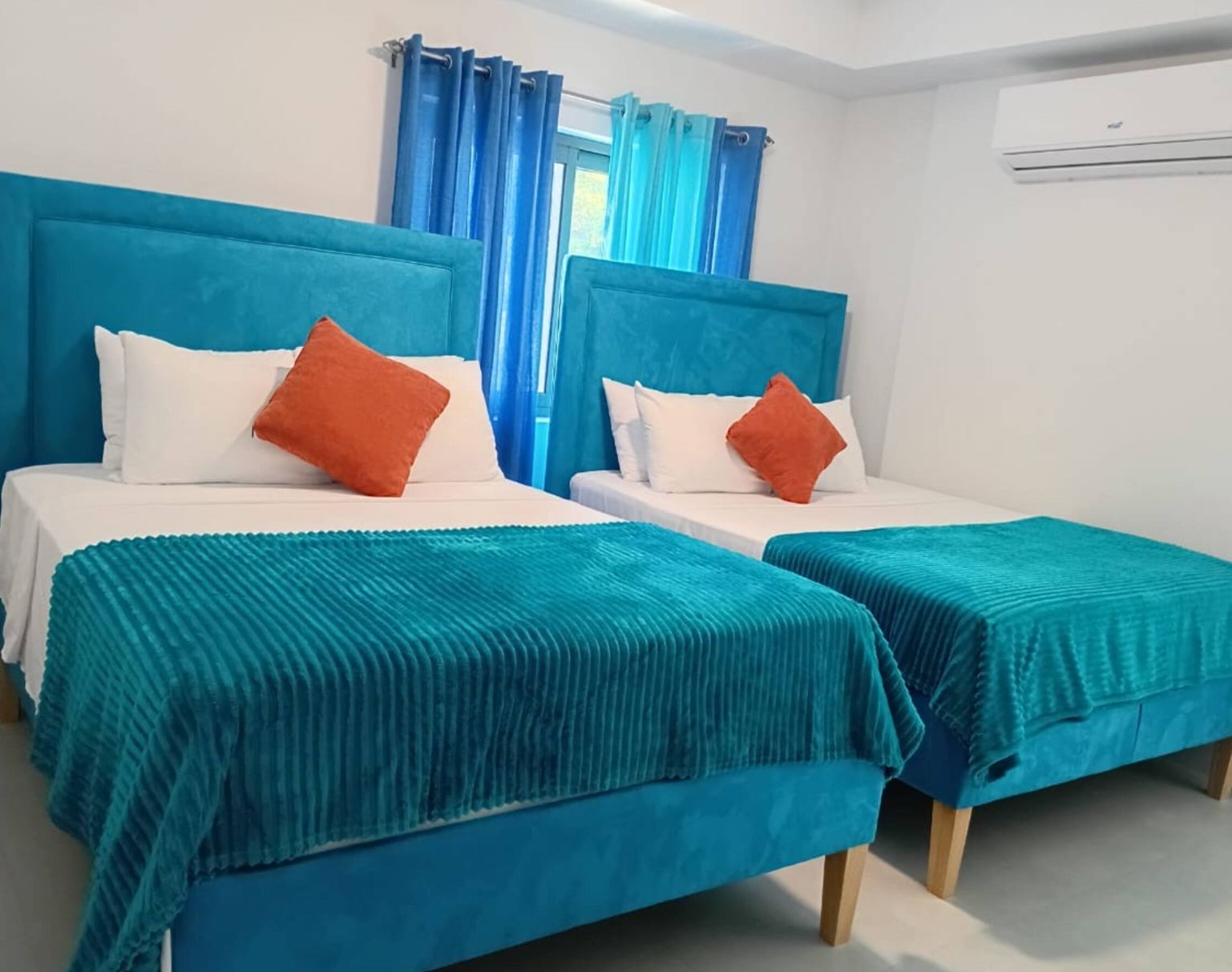 A hotel room with two beds, each with a blue headboard, white bedding, and orange pillows. There is a window with blue curtains, and an air conditioning unit on the wall.