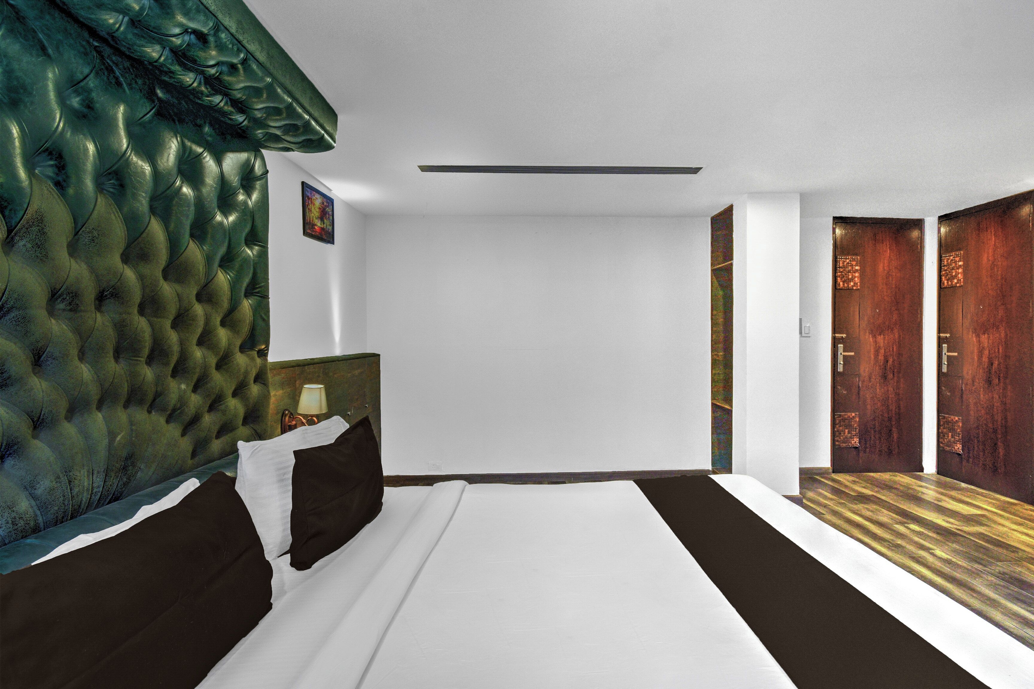 Una habitación de hotel con cabecera capitonada, cama con ropa de cama blanca y oscura, mesita de noche con lámpara y puerta de madera.