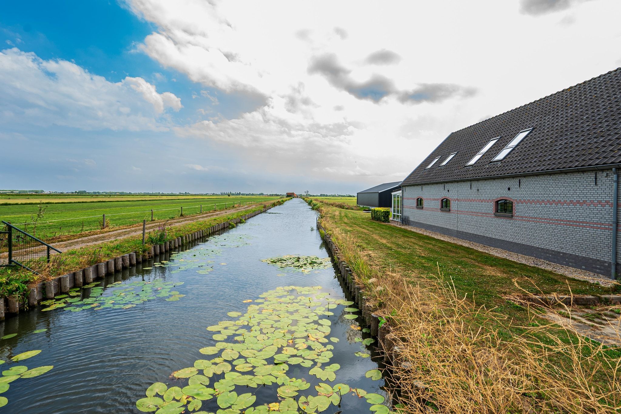Un canale con ninfee, un edificio in mattoni con tetto spiovente e un campo verde sullo sfondo.