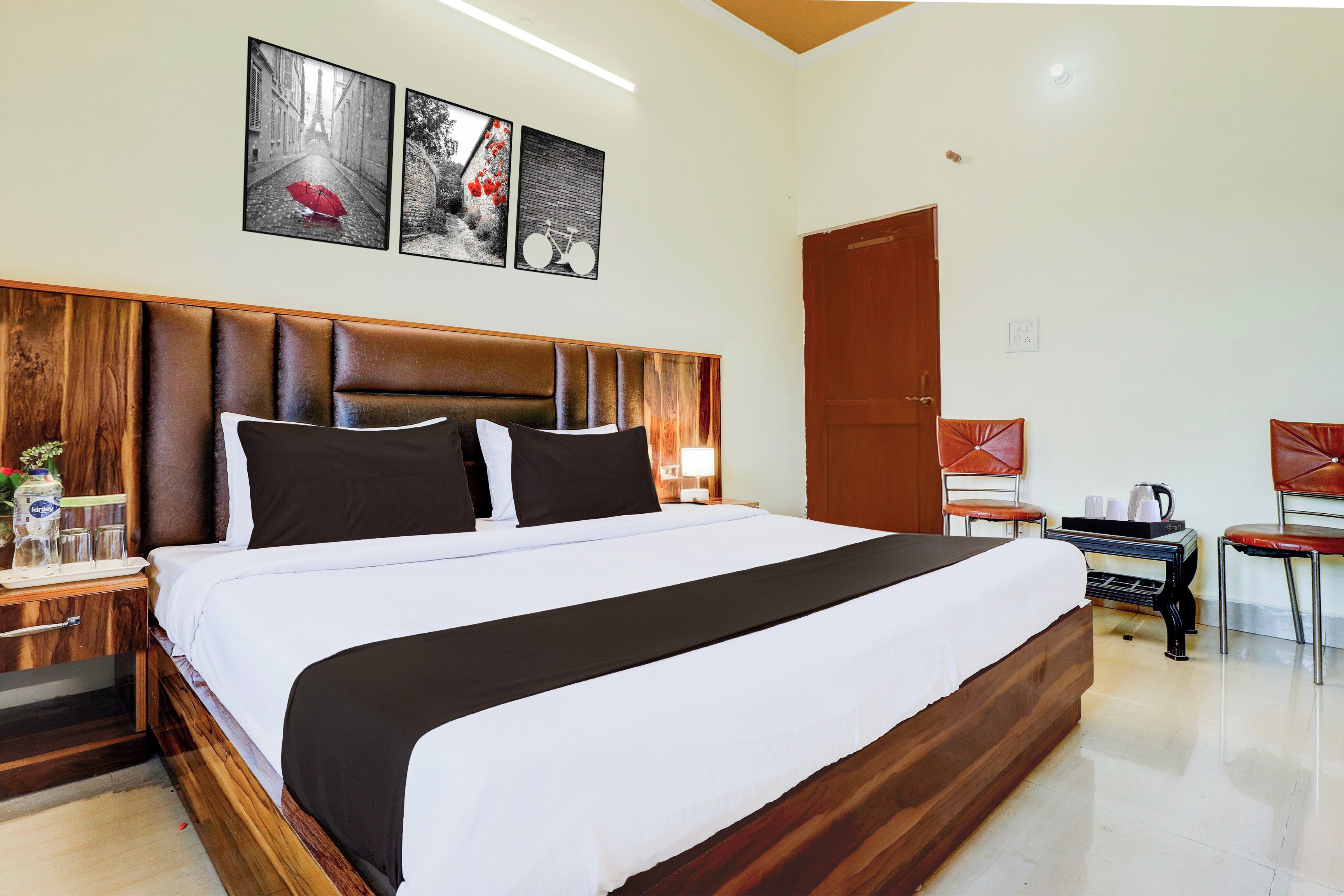A hotel room with a large bed, wooden headboard, a nightstand with a lamp, a wooden dresser, and a seating area with a chair and table.