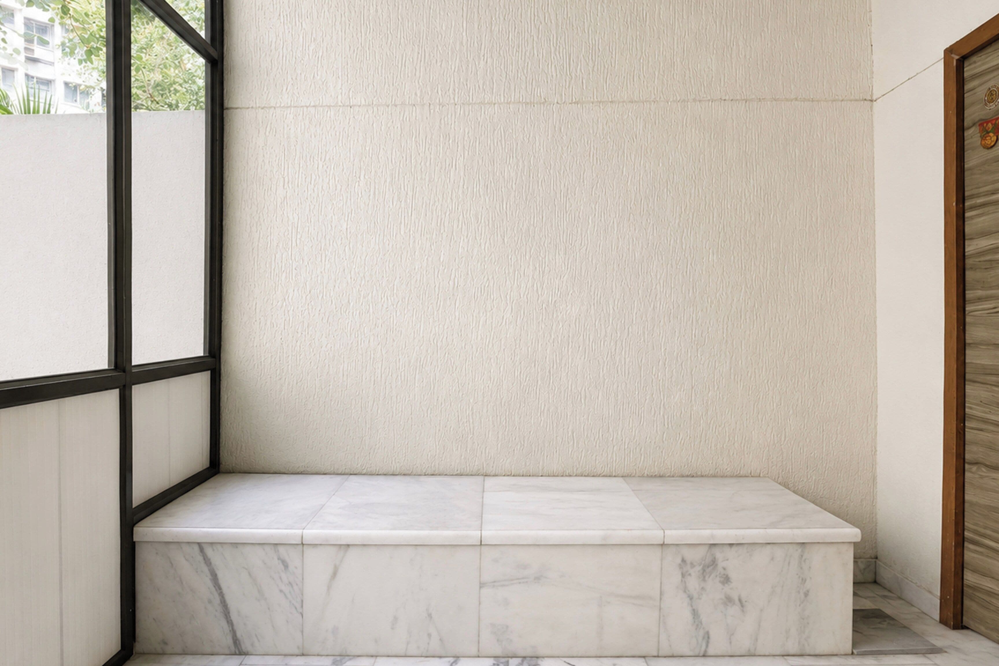 Una esquina de habitación con un banco de mármol, una pared con textura y una puerta de madera.