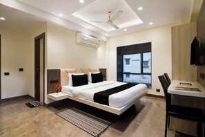 A modern hotel room with a large bed, a desk, a chair, a ceiling fan, and a window with a view of buildings. - Townhouse Newtown DLF (Barasat)