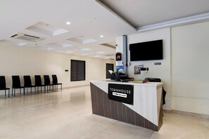 A reception desk with a sign indicating it is serviced by OVO, a waiting area with chairs, a flat-screen TV mounted on the wall, and a security camera. - Townhouse Newtown DLF (Barasat)